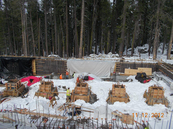 Snow-covered construction site in a forest, with workers and formwork structures.