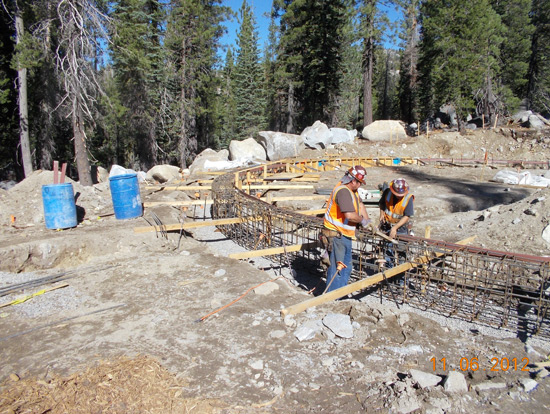 Two workers in safety gear are constructing a curved structure with rebar, surrounded by trees and blue barrels.