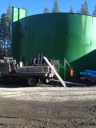 A large green industrial tank with construction materials and equipment nearby, surrounded by trees.