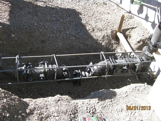 A buried pipe with fittings and valves is exposed in a trench, reinforced with metal bars on a construction site.