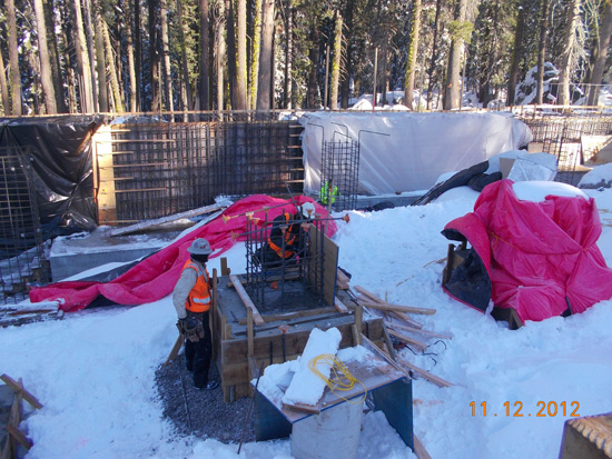 A construction site in a snowy forest with a worker in an orange vest. Pink tarps cover some materials.