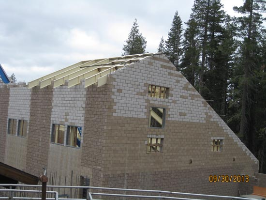A partially constructed building with a wooden roof frame and brick walls, surrounded by trees.