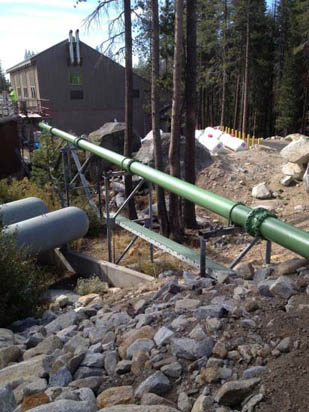 A long, green pipe supported by metal stands runs through a rocky, wooded landscape near a building.