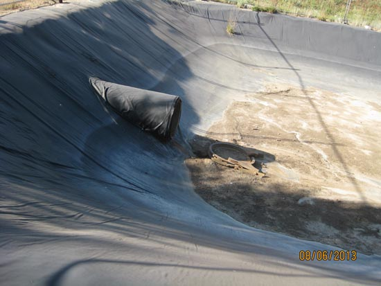 A partially-lined, empty pit with a large black pipe and some dried debris on the ground.