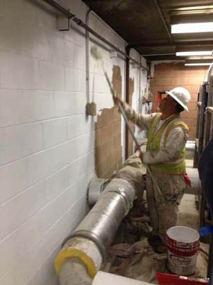 A construction worker paints a wall white in an industrial setting, wearing safety gear including a hard hat and reflective vest.