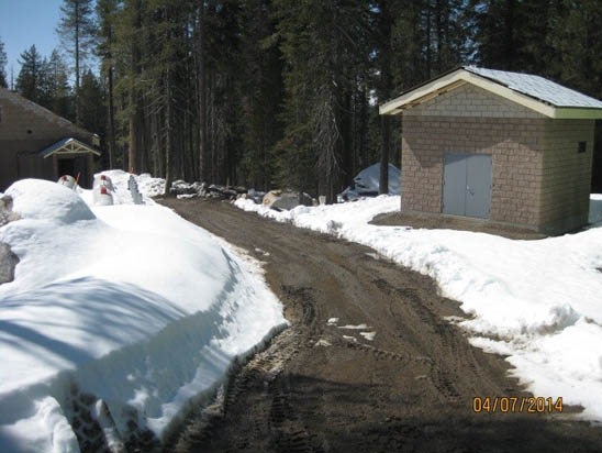 Snowy path through trees, flanked by cabins, with a small building on the right.