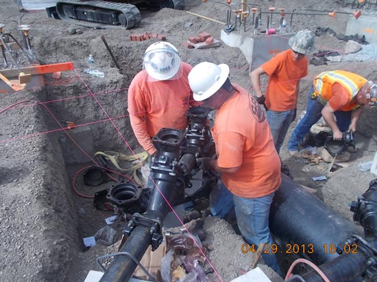 Workers in hard hats and orange shirts installing pipes in a construction site trench.