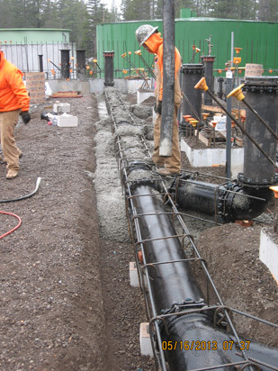 Construction site with workers, pipes, and rebar. Concrete being poured into a trench.