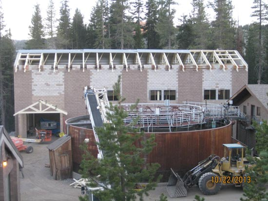 Construction site with a partially built structure and a large cylindrical tank surrounded by trees.