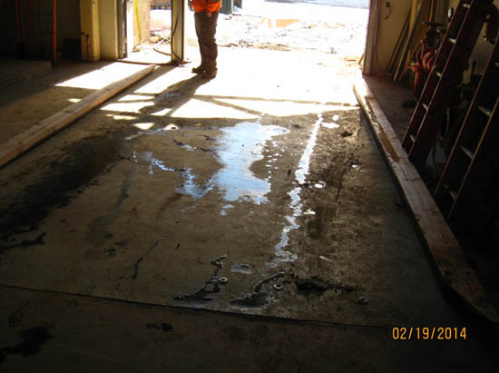 A dimly lit garage floor with wet patches, a person in work attire stands near the bright doorway.