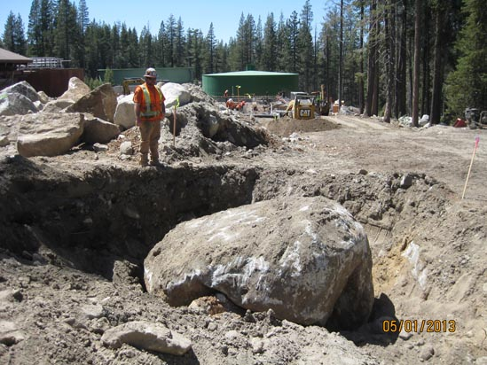 Construction worker standing near a large boulder in an excavation site, surrounded by trees and construction equipment.
