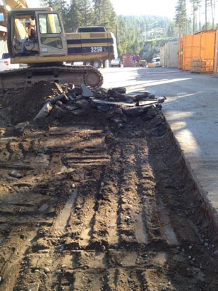 A construction site with an excavator, dug-up dirt, and debris, alongside a partially excavated road or path.
