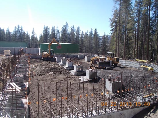 Construction site with heavy machinery, rebar, and concrete foundations surrounded by trees.