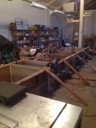 A workshop with shelves of tools and materials, and workstations under construction. A person in a hard hat is working in the space.