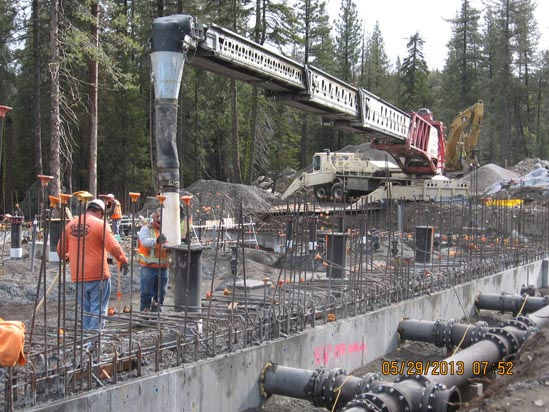 Construction site with workers and machinery pouring concrete, surrounded by trees.