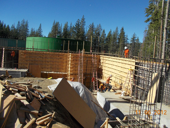 Construction site with workers, wooden frameworks, rebar, and a large green tank in a forested area under a clear blue sky.