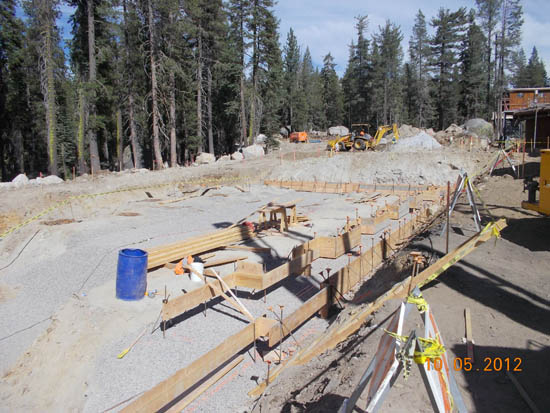 A construction site in a forest clearing with wooden frames, a blue barrel, and machinery in the background.