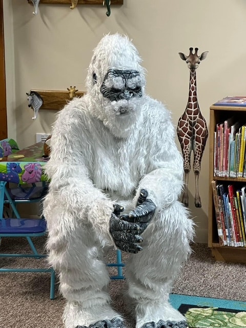 A person in a furry white Yeti costume sitting in a children's area, surrounded by books and a giraffe mural.