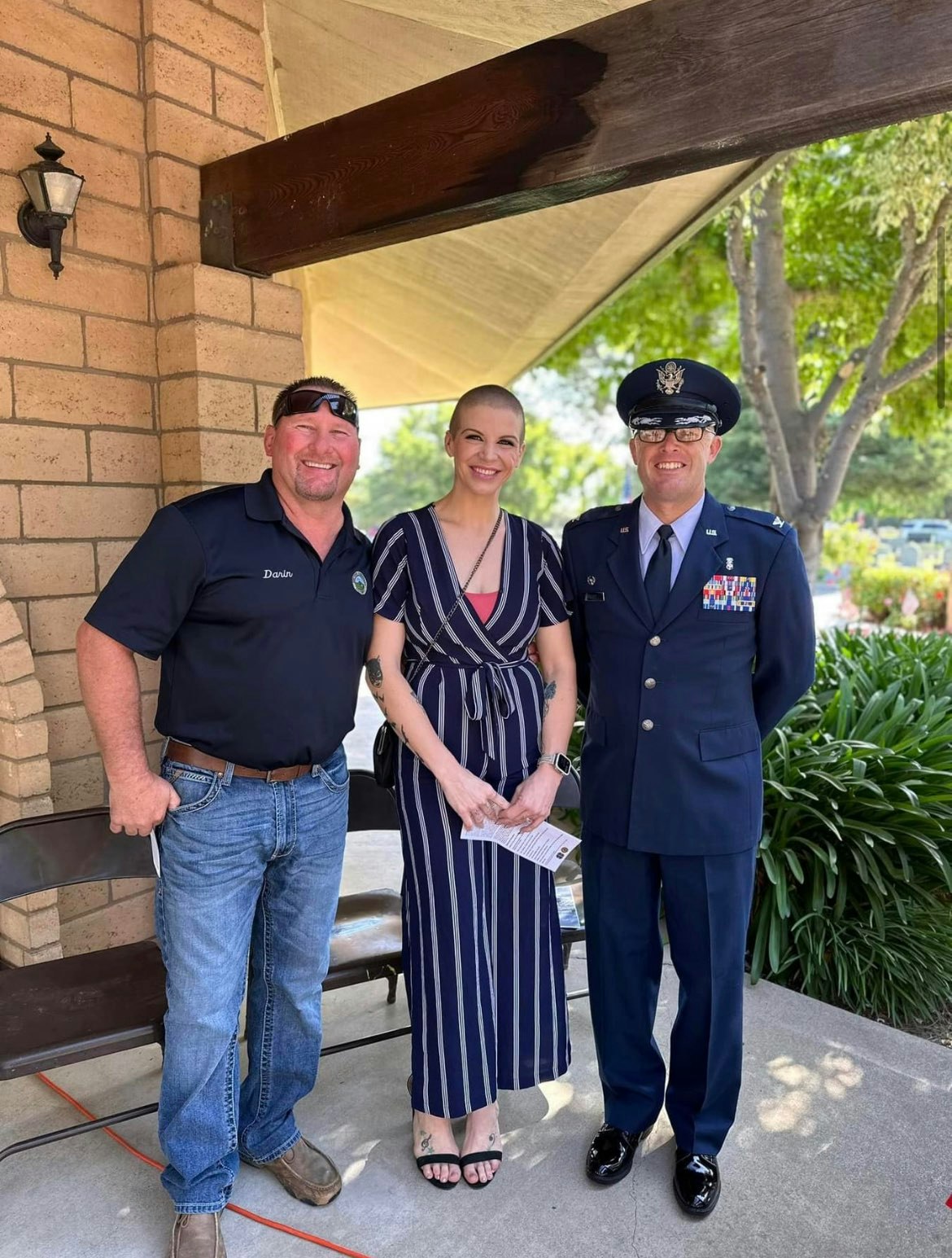Three people stand together outdoors, one in military uniform, another in casual attire, and a woman in a striped dress.