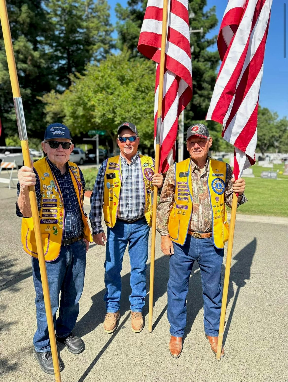 Three older men stand together holding American flags, wearing yellow vests with various badges, in a green outdoor setting.