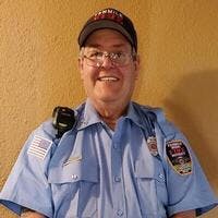 An older man wearing a blue uniform with a badge and a cap, smiling for the camera.