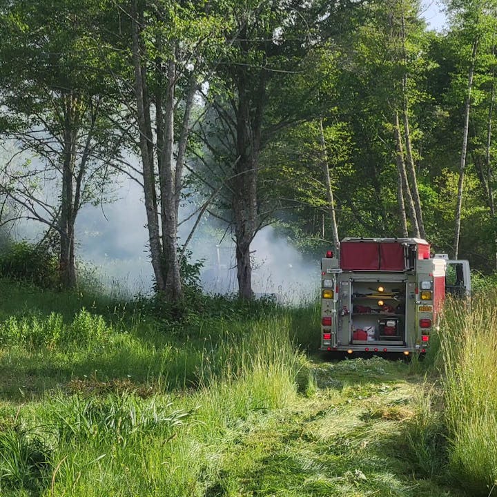 A fire truck is positioned in a grassy area, with smoke visible among the trees, indicating a potential fire nearby.