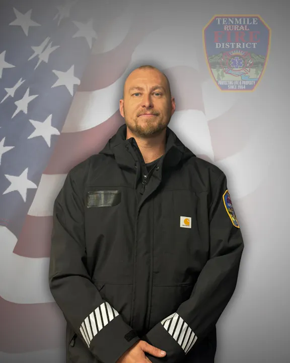 A person in a black jacket stands before an American flag backdrop with a "Tenmile Rural Fire District" badge.