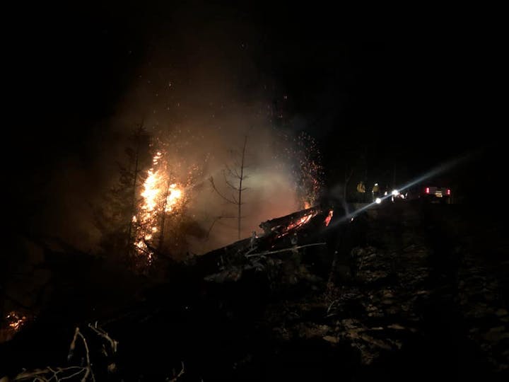 A nighttime scene shows a wildfire burning trees with smoke and sparks, alongside emergency response vehicles in the background.