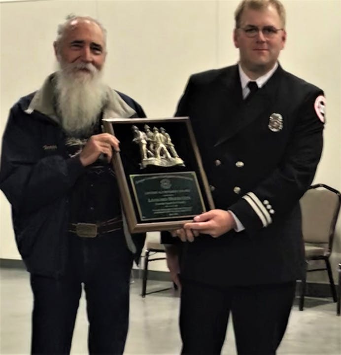 Two men, one with a beard and the other in a firefighter uniform, are holding an award plaque together.