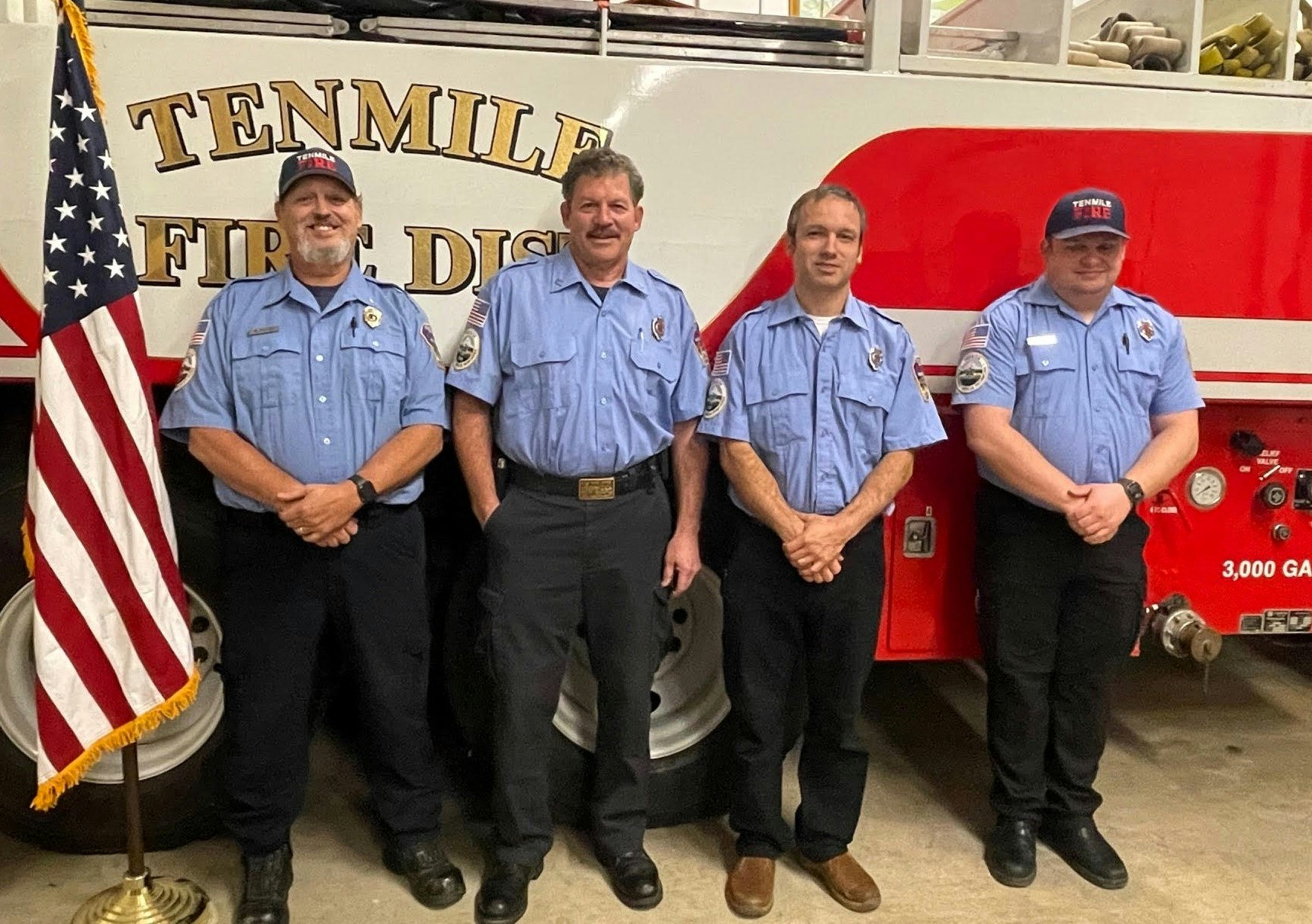 Four firefighters in blue uniforms stand in front of a red fire truck, alongside an American flag, representing Tenmile Fire District.