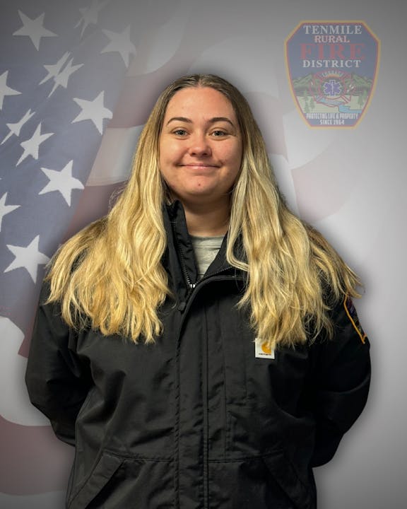A person in a black jacket stands in front of an American flag and a "Tenmile Rural Fire District" emblem.