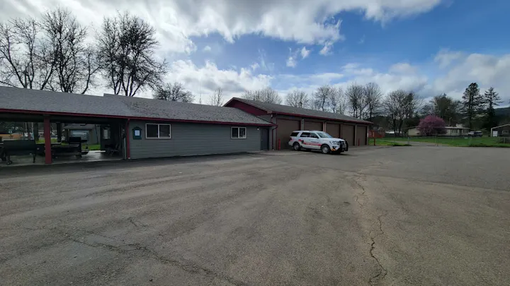 The image shows a building with a gray exterior, a parked emergency vehicle, and trees under a cloudy sky.