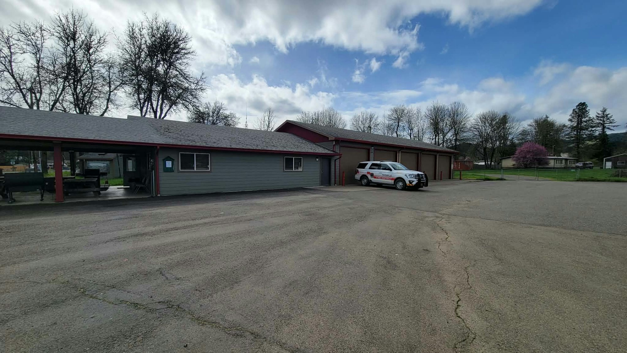 The image shows a building with a gray exterior, a parked emergency vehicle, and trees under a cloudy sky.