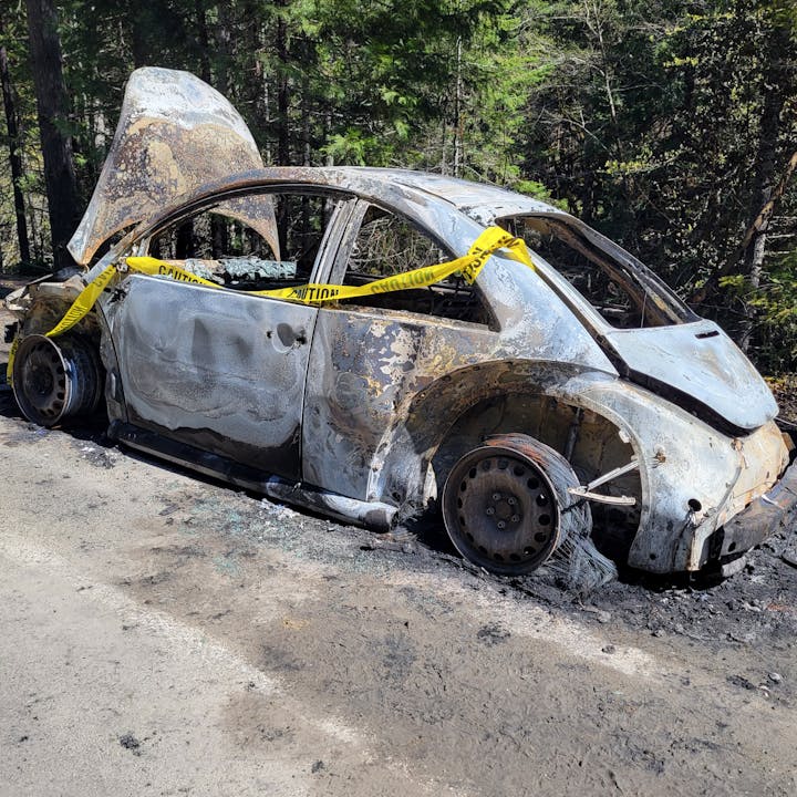 Burnt-out car with caution tape around, on a road by a forest.
