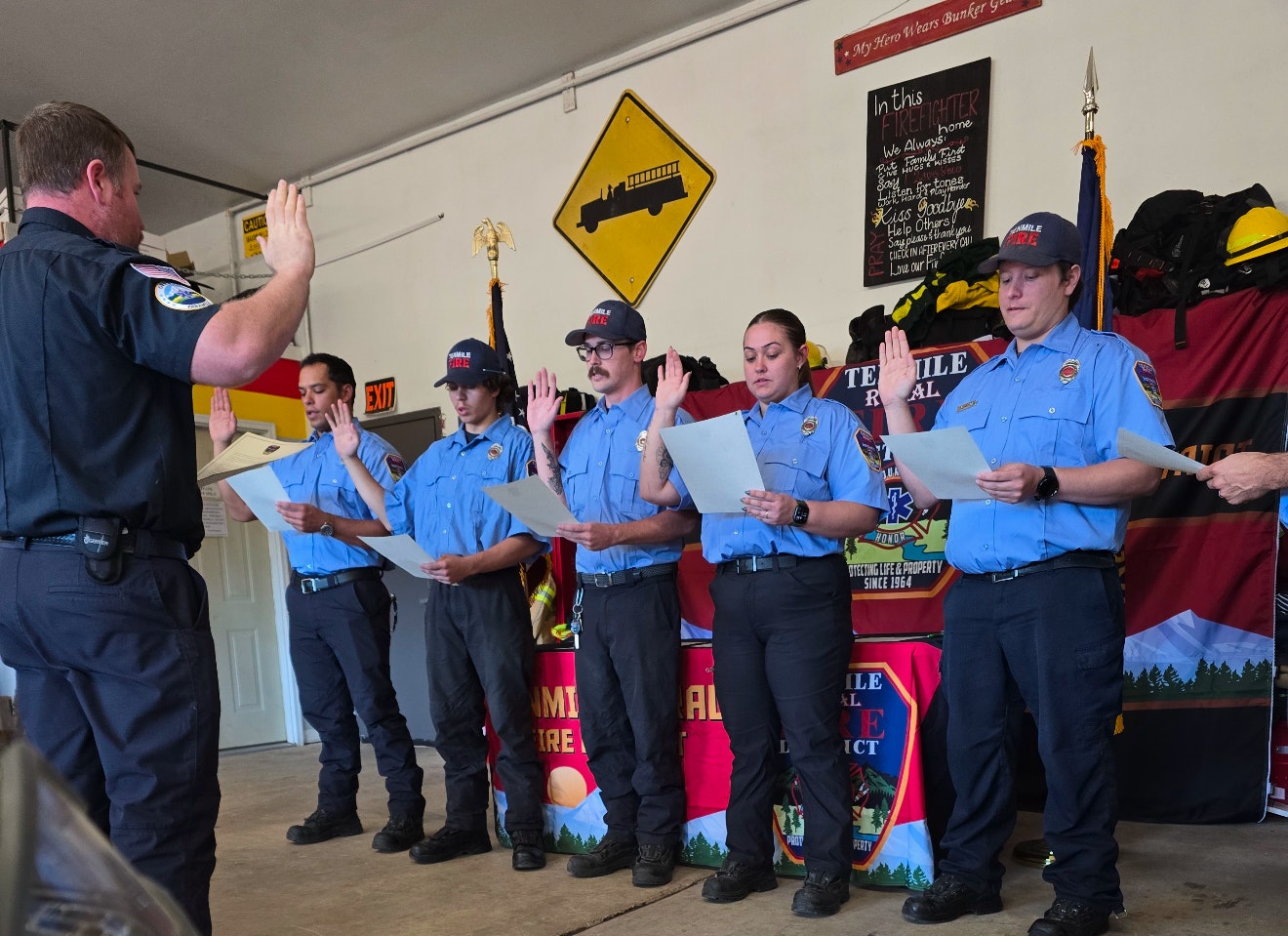 A group of firefighters taking their oath in a ceremony, led by an officer, in a fire station setting.