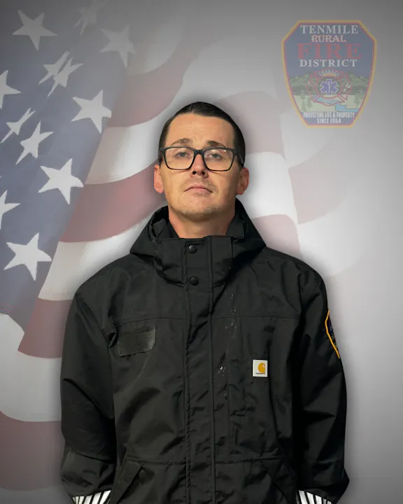 A person in a black jacket stands in front of an American flag and a fire district badge.