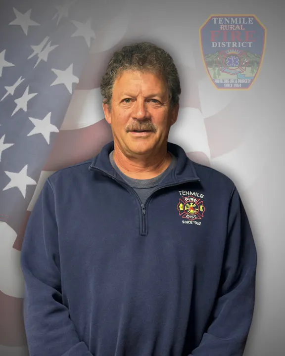 A man in a blue sweatshirt with a fire department logo, standing before an American flag backdrop.