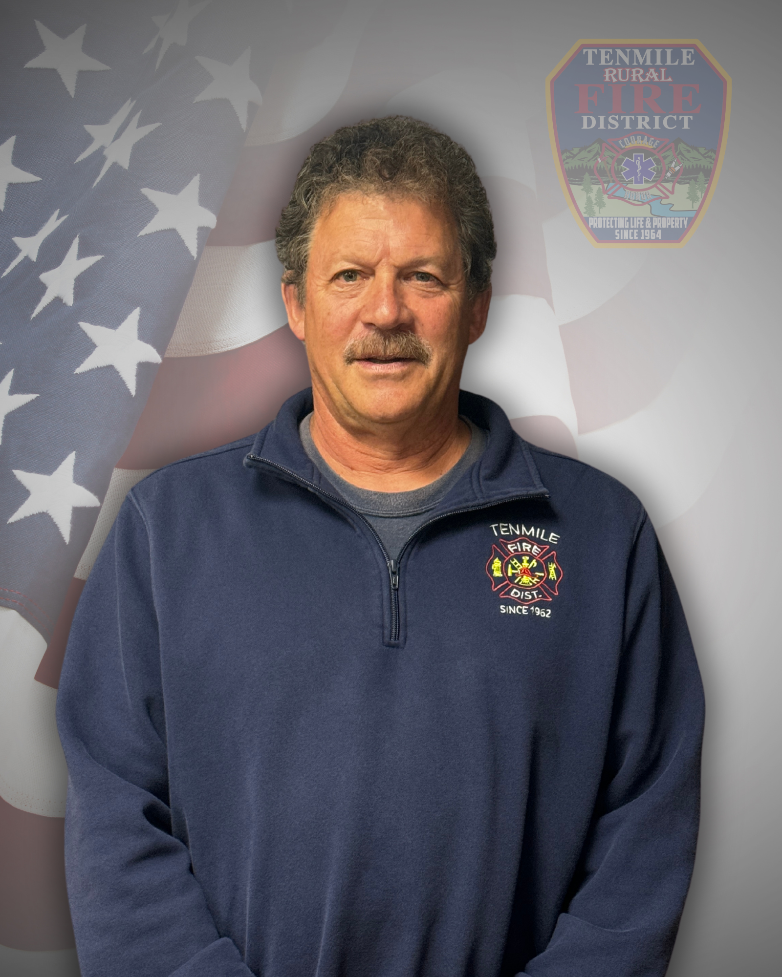 A man in a blue sweatshirt with a fire department logo, standing before an American flag backdrop.