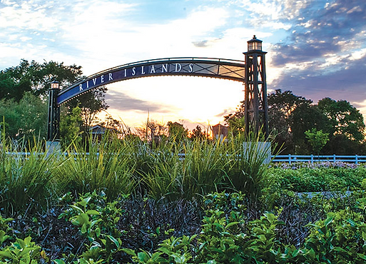 Entrance arch with "River Islands" sign, plants in foreground, sunset sky in the background.