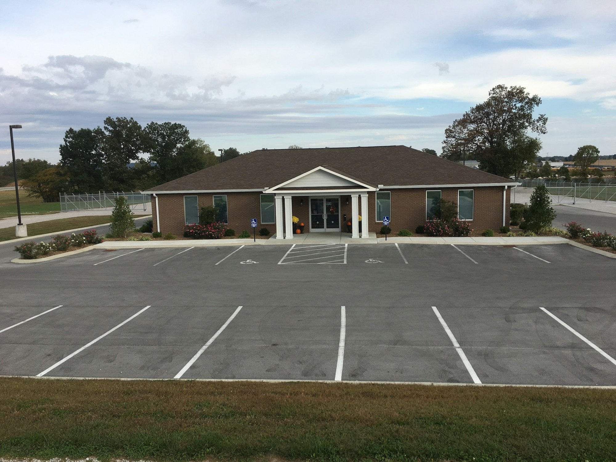 A small office building with a gabled roof, surrounded by an empty parking lot and landscaped with plants and trees.
