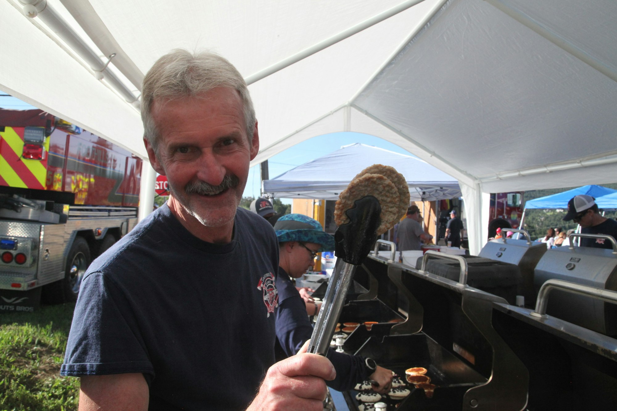 A man holding a sausage with tongs, standing by a grill at an outdoor event.