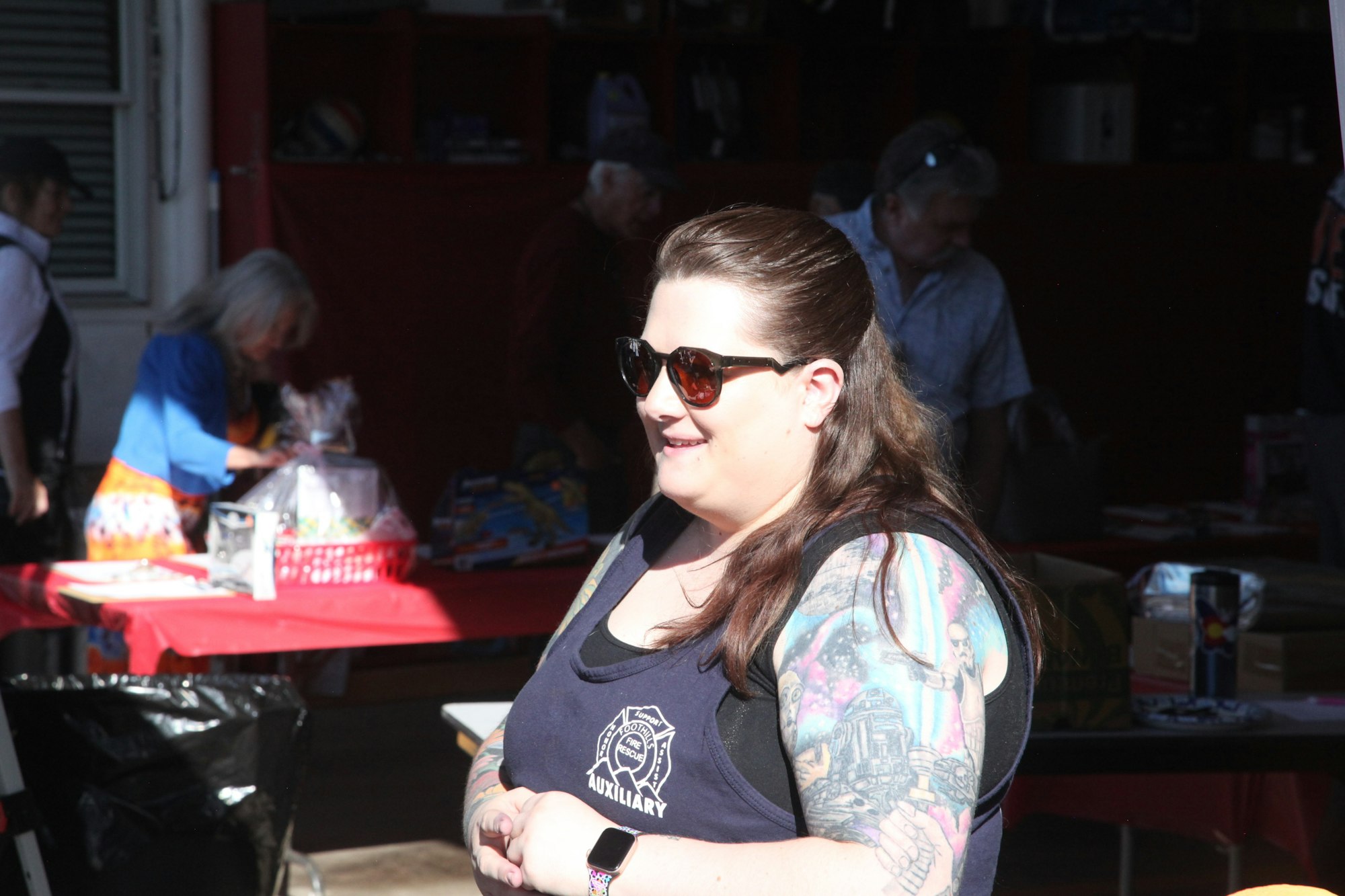 A woman wearing sunglasses and a tattooed arm is smiling outdoors; people and a table with items are in the background.