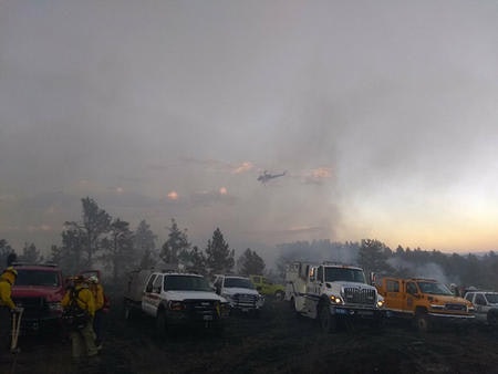 Firefighting vehicles and personnel in a smoky area; a helicopter flies above.