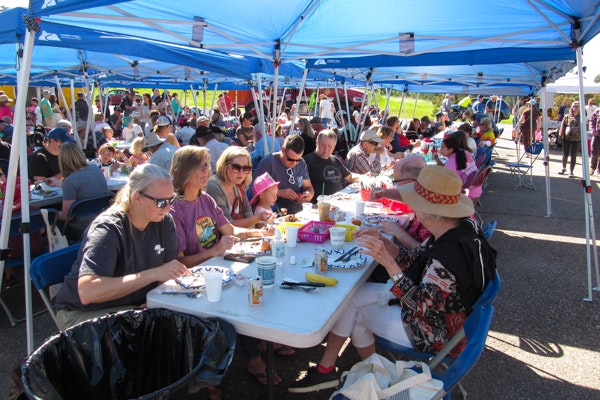People are sitting under blue canopy tents, eating and socializing at a large outdoor gathering.