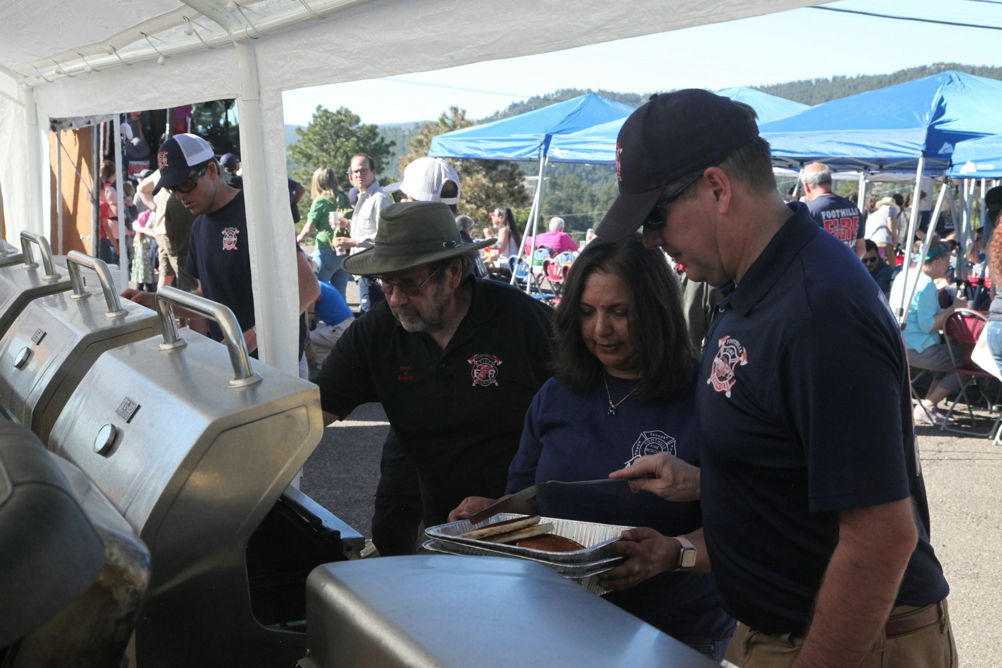 People cooking and serving food at an outdoor event, with grills under a tent.