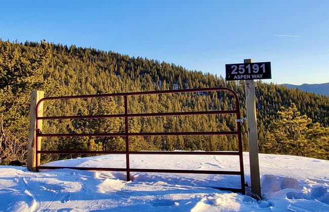 A snowy landscape with a red gate marked "25191 Aspen Way" and a forested hillside in the background under a clear blue sky.
