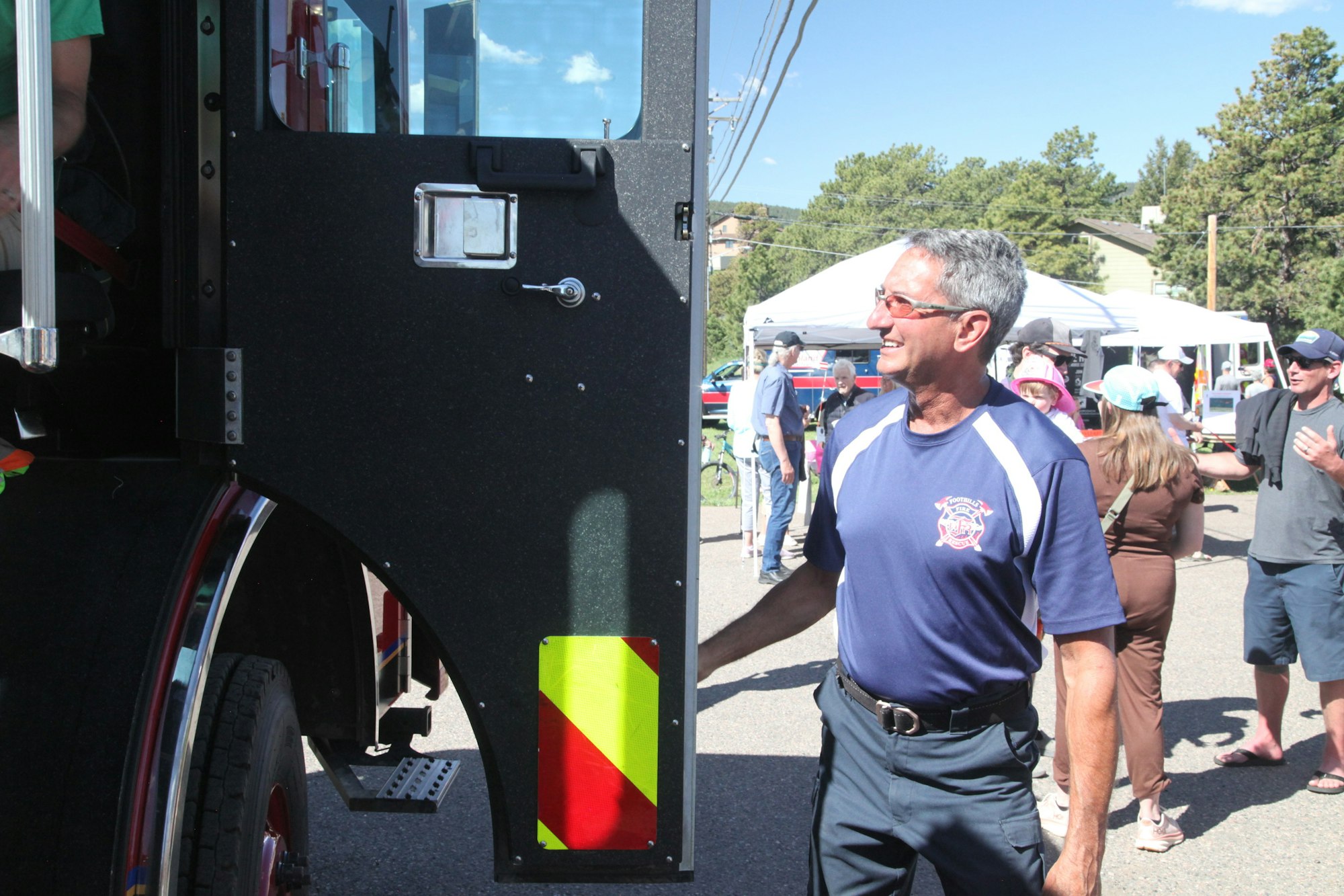 A man in a firefighter shirt stands beside an open fire truck door. People and tents are in the background.