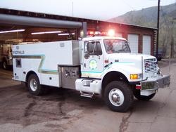 A white firefighting truck with lights on, parked outside a garage.