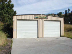 A metal garage with two closed doors and a sign on top, surrounded by grass and trees.