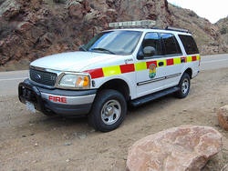 A white fire service SUV with red and yellow markings is parked on a roadside near rocky terrain.